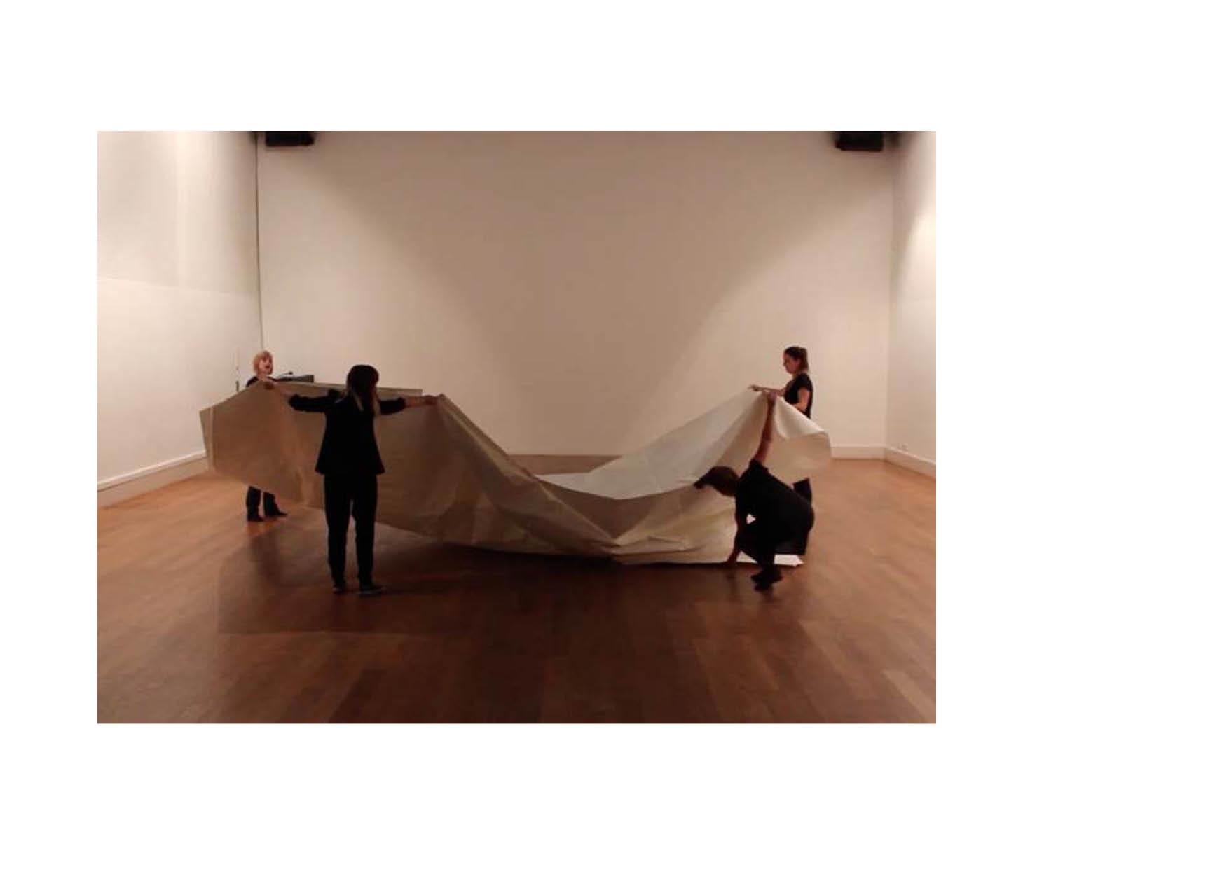 Four people that seem to hold a big, creme-white paper in a light space with wooden floor. Twee people stand on the right side one holding a cornern , one underneed the under the sheet of paper. The sheet of paper is formed in a wave like form.